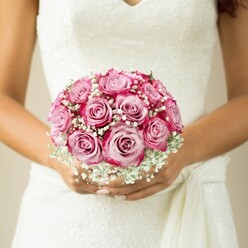 In the serene light of a New Barnet morning, a bride's hands cradle a flawless, hand-tied bouquet - a testament to refined artistry and local floral tradition. Each rose in the arrangement displays a captivating transition: petals swirl from a blissful, pale pink heart toward edges saturated in bold magenta, revealing a depth of colour only rivalled by the joy of the occasion. Wisps of baby's breath, so often seen in the finest North London arrangements, nestle between the full blooms, creating an airy, cloud-like halo that softens the bouquet's vivid palette. The bride's skin glows warmly against the delicate floral interplay, while her perfectly manicured nails brush gently against the silk wrap around the stems. Her wedding gown's lace bodice, with gentle floral motifs and a graceful V-shaped neckline, adds traditional charm, the fabric pooling softly just above her hands. The muted, neutral background ensures every detail of the bouquet and the bride's poised embrace remains the centre of focus. Throughout the scene wafts an imagined scent of dewy roses and sweet gypsophila, evoking both romance and anticipation. For couples marrying in New Barnet or the leafy surrounds of East Barnet and Hadley Common, this bouquet encapsulates the emotion, elegance, and sense of cherished place that define a truly unforgettable day.