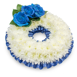 Seen from a modestly elevated viewpoint against a pure white surface, this circular sympathy wreath presents itself with calm formality and gentle texture. The central composition is a full, uninterrupted band of white chrysanthemums; each bloom contributes soft, feathered layers, creating a slightly domed, cloud-like effect whose subtle variations in petal and centre tone read as tactile warmth. On the upper left, three vivid blue roses rise as a striking focal cluster - their petals dense and velvety, coloured in a deep royal-cobalt that feels both solemn and serenely beautiful. Around the roses, deep green foliage sits low and glossy, its rounded leaves offering a natural cushion that enhances the roses' prominence without overpowering the chrysanthemum field. A finely pleated satin ribbon in the same blue hues borders the wreath's outer and inner rims, its gentle ripples and reflective surface bringing visual rhythm and a finished, ceremonial edge to the arrangement. The soft shadow beneath the wreath anchors it to the surface, while even, diffused light reveals the delicate textures and the floral craftsmanship. This wreath reads as an understated yet noble expression of condolence - a carefully composed memorial piece meant to convey comfort, respect, and lasting memory. As someone who works locally around New Barnet and Brunswick Park, I often recommend this palette when families request a traditional, heartfelt tribute that balances purity with a personal, calming touch.