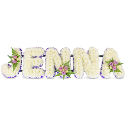 This intimate, bespoke tribute presents the name JENNA in large, sculpted letters, each formed from a dense mass of snow-white chrysanthemums or similar small, rounded florets that give the piece a cloud-like, comforting volume. The letters sit against a clean white background so every petal and contour is visible; a delicate border of deep purple ribbon or tiny lavender florets traces the inner and base edges of each character, giving crisp definition and a soft regal note. To personalise the composition, three small accent sprays are artfully placed - on the upper curve of the J, at the center bottom of the second N, and on the upper right of the A - each composed of blush-pink and pale lavender spray roses, interspersed with cream or pale yellow rosebuds and fine fern fronds that lend a fresh green scent and feathery texture. Subtle dark connecting bars beneath the letters ensure structural unity without drawing attention away from the blooms. The lighting is gentle, suggestive of a quiet morning or late-afternoon service, letting shadows fall softly between petals and highlighting the petals' satin sheen. As a sympathy arrangement, the design balances purity and tenderness: the white chrysanthemums convey serenity, the purple trim offers dignity, and the pastel rose clusters intimate warm remembrance. Locally, this kind of personalised floral name tribute is a respectful choice for wakes or services held around New Barnet, on Station Road or near the High Street - a carefully made, deeply felt expression of condolence and memory.