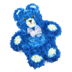 Set against a clean, white studio backdrop, a floral teddy bear stretches out in a calm, reclining pose, its form meticulously built from a carpet of royal blue petals. The dense cluster of small flowers, reminiscent of chrysanthemums or similar button blooms, covers the entire body, from the rounded head down to the plump feet, creating the illusion of soft fur you could almost stroke. The florist has shaped every curve with care: the outline of the arms resting by the sides, the gentle slope of the belly, and the slightly tilted head that lends the bear a tender, almost listening expression. Cream-toned flowers, edged with the faintest hint of yellow, define the snout, inner ears, and the oval pads on each paw, giving the face and limbs a friendly clarity against the deeper blue. Two shiny, dark brown eyes, set close enough to feel endearing, catch the light with a subtle gleam, while a small black triangle nose and a fine curved line below it form a soft, reassuring smile. Around the neck sits a sheer white bow, almost like organza, its airy folds resting lightly on the chest and suggesting both ceremony and affection. The overall composition feels peaceful and thoughtfully balanced, ideal as a sympathy tribute or memorial design that might be delivered to local chapels, homes, or crematoria around New Barnet and nearby East Barnet or Oakleigh Park. As a florist familiar with the community, I see this piece as a floral companion for moments of farewell or remembrance, especially for a baby boy or young child, offering quiet comfort in its colour, texture, and gentle presence.