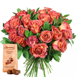 As a seasoned New Barnet florist, I see this radiant bouquet and gourmet chocolate pairing as a celebration of life's sweetest moments, imbued with care and artistry. Gathered with precision, each rose opens in a gentle, swirling cascade-from coral hearts flaring outward to delicate bands of peach and salmon, finishing in blushes of rich pink. Scattered throughout, glossy, verdant greenery frames the flowers without overwhelming them, the contrast reminiscent of the mature tree canopies lining nearby East Barnet Road. The tactile allure is striking-the petals' velvet sheen and soft curves catch the light, inviting both visual and sensory appreciation. Tucked at the bouquet's base is a Thorntons Orange Crisp chocolate pouch, its creamy, understated design offering a quiet counterpart to the exuberant blossoms. The handwritten script and tempting illustration, along with two glossy, unwrapped milk chocolate truffles, evoke a sense of shared indulgence. This artful composition, perfect for gifting on special occasions or simply uplifting a loved one's day, transports the senses to a realm of fragrant blooms and sweet treats, rooted firmly in the welcoming, familiar atmosphere of New Barnet.