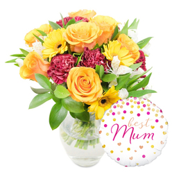 This image presents a thoughtfully arranged, celebratory bouquet in a simple clear glass vase, photographed on a pristine white background so every colour reads clearly. At the heart are several peachy-orange and golden-yellow roses, their petals thick and velvety, radiating a soft, honeyed scent that seems almost visible. Sunny yellow gerbera daisies peek between the roses, their bold petals and dark centers adding joyful punctuation, while deep burgundy carnations bring a plush, textured counterpoint with their densely ruffled edges. Interspersed throughout are crisp white alstroemeria - Peruvian lilies with narrow, speckled petals - which introduce delicate patterning and a lighter fragrance. The flowers sit within a generous embrace of mixed greenery: broad glossy leaves that catch the light and finer sprigs that add movement and freshness. The glass vase shows neatly trimmed stems and clear water, a small detail that signals careful floristry. Nestled to the bottom right and overlapping the vase is a round white foil balloon decorated with scattered gold and fuchsia dots and little hearts; the central message "best Mum" is written in a flowing hot-pink script, adding an undeniably personal, celebratory touch. The mood is sunny and affectionate, ideal for marking Mother's Day, a special birthday, or any moment of thanks. As a local florist would note, this arrangement suits deliveries across streets such as East Barnet Road and around Hadley Green - a familiar, heartwarming choice for neighbours in New Barnet seeking to send something bright and sincere.