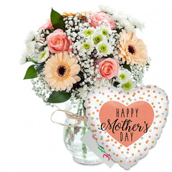 Against a clean, white studio-style background, a Mother's Day floral gift arrangement glows with soft colour and heartfelt sentiment, as if it has just been prepared in a New Barnet flower shop on a calm spring morning. The eye is first drawn to the generous bouquet arranged in a clear glass vase, its simple cylindrical form wrapped at the neck with a casual bow of natural twine that hints at a homely, handmade touch. Inside, several peach-pink roses are fully open, their petals spiralling outward in velvety layers that suggest gentle fragrance and tenderness. Alongside them, large peach gerbera daisies with dark, almost chocolate-brown centres create a bright, sunny contrast, echoing the cheerful front gardens you might see on a walk from New Barnet station towards East Barnet Village. Interwoven among these focal blooms are airy sprigs of white baby's breath, each tiny flower a dot of softness, and neat white chrysanthemum heads topped with light green centres, giving the arrangement structure and volume. Rich green foliage frames the blooms, some leaves glossy, others more matte, all contributing to a sense of natural abundance and freshness. Nestled behind the bouquet, just visible between stems and balloon, is a small white card with colourful, slightly blurred writing that suggests a personal Happy Mother's Day note. In front of the vase to the right, a heart-shaped foil balloon adds a touch of playful celebration. Its white surface is scattered with metallic gold and silver dots, and at the centre a peach-toned heart holds the message HAPPY Mother's DAY in bold black lettering and flowing script. The composition feels joyful yet tender, ideal for capturing the love and appreciation families across New Barnet wish to send on Mother's Day.