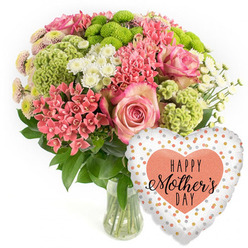 A lavish Mother's Day bouquet is photographed against a bright, uncluttered white backdrop, creating a calm gallery-like setting that lets every blossom stand out, much like a carefully curated display in a New Barnet high street flower shop. The arrangement in the clear glass vase is rounded and generous, brimming with texture and colour. Blush-pink roses, with their deeper pink edges and spiralling centres, offer an immediate sense of tenderness, as if chosen to say thank you after a family lunch near New Barnet Station. Around them, small coral-pink blossoms, similar to kalanchoe or clustered spray flowers, weave through the design, introducing a lively, almost sparkling warmth. Vibrant lime-green button chrysanthemums and velvety, wavy chartreuse florets add a bold, contemporary twist, their sculptural shapes inviting you to imagine their firm, almost cushion-like feel beneath your fingertips. Soft white daisy-style spray chrysanthemums scatter light throughout the bouquet, their simple petals giving the eye a place to rest amid the richer hues. Deep green leaves nestle closely around the stems, grounding the brighter tones and suggesting freshly cut foliage straight from a trusted local supplier. The vase itself is clear and understated, revealing the straight stems and clean water that keep the flowers looking newly arranged. To the right, a metallic heart-shaped balloon adds a distinctly festive accent. Its white surface shimmers with scattered dots of gold, silver, and peach, drawing the gaze toward the larger coral-peach heart in the centre, printed with the phrase HAPPY Mother's DAY in a stylish black script. Together, bouquet and balloon create a joyful, ready-to-deliver gift, perfect for surprising a mum at home in New Barnet, whether she's near Victoria Park, East Barnet Village, or tucked away on a quiet residential street.