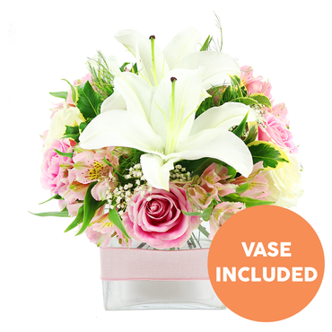 A refined floral centerpiece is photographed against a bright white background, its compact, sculpted form giving an immediate sense of calm elegance. Dominating the middle of the design, pristine white Oriental lilies open wide, their smooth, waxy petals arching gracefully and exposing rich reddish-brown stamens that add a hint of drama. Encircling these lilies, layers of blush and soft pink roses create a gentle halo, each bloom at a slightly different stage, from tight buds with curled edges to fully open flowers with velvety, ruffled petals. Between them, you can trace the slender forms of pink and cream-white alstroemeria, some petals decorated with fine yellow stripes and freckles, adding an intricate, almost hand-painted detail. Airy tufts of white baby's breath are interspersed throughout, lightening the composition so it feels as though parts of the bouquet are floating. Rich greenery in multiple shapes and sizes - glossy leaves, narrow blades, and delicate fern-like sprigs - frame the flowers and provide a fresh contrast, similar to the lush hedges and tree lines you might notice along New Barnet's quieter residential streets. All of this is presented in a clear rectangular glass vase, the stems cleanly aligned and visible through the water, with a narrow pale pink ribbon wrapped neatly around its middle for a soft, romantic finish. In the bottom right corner, a bright orange circle with bold white capitals clearly states VASE INCLUDED, making it obvious that the container arrives as part of the gift. The bouquet exudes a mood of tranquillity and gentle joy, perfectly suited to a bedside table, a dining nook, or a thoughtful same-day flower delivery in New Barnet.