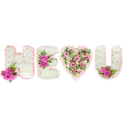 In soft studio light, this bespoke sympathy tribute spells out H, E, a heart, and U in a layout that reads like a handwritten vow in flowers. The H and E are meticulously formed from compact white chrysanthemums; their tiny petals create a velvety, cushion-like surface that calms the eye. Each letter is edged in a light blush satin ribbon with a delicate ruffled finish, a feminine, comforting frame. Accents of vivid fuchsia roses provide dramatic contrast: a rich pair nestled among small red berries and deep green, ferny foliage brightens the base of the H, while a bunched trio of the same roses anchors the horizontal bar of the E, their aroma adding a warm, familiar sweetness to the composition. Center stage is a fully rounded heart, composed of layered soft-pink carnations, pale lisianthus and small rosy spray blooms, interlaced with pale green eucalyptus that softens the pinks and introduces a subtle, minty fragrance. The U repeats the white-chrysanthemum construction and blush ribbon border, finished with a small cluster of fuchsia roses and foliage tucked into its inside curve for symmetry. As a New Barnet florist would make for a local memorial - whether to place by New Barnet Station or at a family home near Victoria Recreation Ground - this design balances dignity and affection. Prepared with attentive technique and respectful intent, the display reads as both a declaration of love and a tender remembrance, appropriate for funeral services, commemorative gatherings, or as a quiet, heartfelt message from neighbours and friends. (Flowers New Barnet can arrange delivery across New Barnet and surrounding streets.)