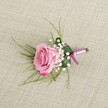 A delicately crafted pink rose boutonniere rests on a soft, textured beige linen cloth, captured in gentle, even light that feels like a calm morning in New Barnet. Viewed from a slightly elevated, eye-level angle, the central rose is a semi-open blush pink bloom, its velvety petals swirling outward in tight spirals from a pale, almost shell-pink heart to edges that deepen into a richer, romantic magenta. Around the base of this single rose, clusters of creamy white baby's breath create a halo of tiny, cloud-like blossoms, each floret no bigger than a pinhead yet together forming a soft, airy cushion that enhances the rose's fullness. Wisps of slender, grass-like greenery arc out from behind, their bright, fresh green tones adding movement and a sense of natural lightness, reminiscent of the greenery you might see along the paths in nearby Oak Hill Park. A broader, darker green leaf is folded neatly at the base, cupping the stems and lending structure and polish, much like a tailored lapel on a wedding suit. The stems are carefully bound with a lustrous pink satin ribbon, tied into a precise, modest bow whose short tails lie smoothly against the linen, echoing the rose's blush tones without overwhelming them. A slim, silver-toned pin peeks discreetly from the base, ready to secure the piece to a groom's or prom guest's jacket for a celebration in or around New Barnet. The woven threads of the linen backdrop are subtly visible, adding a natural, handcrafted feel that mirrors the attentive florist's touch and the quiet sophistication of a classic, romantic boutonniere designed for a day filled with affection and ceremony.