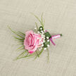 A romantic pink rose boutonniere lies on a pale beige, linen-like fabric, its position slightly angled as if just placed down while getting ready for a wedding in New Barnet. The rose is the undeniable focus: a lush bloom whose petals show a smooth gradient from deep fuchsia at the tips to a soft, powdery pink toward the center, each layer unfolding in a gently cupped spiral. Around the rose, tiny clusters of crisp white baby's breath create an airy frame, their small, star-like blossoms bringing a sense of purity and celebration. Wispy, grass-like green foliage fans out from beneath and behind the rose, adding length, movement, and a fresh garden feel that might remind one of a quiet walk through the leafy streets off East Barnet Road. A single, broader emerald green leaf is curled protectively at the base, its surface dotted with a few clear water droplets that suggest careful misting just before delivery. The stems are tightly bound with a smooth satin ribbon in a light pink shade, wrapped with precision to form a slim, elegant handle. At the base, a darker magenta-pink tie introduces a subtle two-tone detail, catching the light and accentuating the rose's deeper hues. The neutral background, with its gentle texture, keeps the focus on the interplay of soft pinks, sharp whites, and rich greens. The boutonniere feels personal and thoughtful, ideal for a groom, best man, or prom guest in New Barnet who wants a small but meaningful floral accent to mark an important occasion.