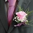 There is a quiet poetry to this New Barnet boutonniere, each element chosen with heartfelt care to capture the spirit of celebration and promise. At its core rests a blush-pink rose, its petals soft as memory, half-unfurled to reveal a gentle vibrance topped with invisible morning dew. The rose is surrounded by sprigs of ethereal baby's breath-each tiny floret a wisp of white light-balancing the central bloom with a halo of cloudlike softness. Slender, textured greens reach out behind, reminiscent of the tranquil gardens near Church Hill Road, lending natural elegance and fresh energy to the composition. Anchoring the piece is a neat bow of lavender satin ribbon, subtle and refined, catching hints of lilac from the light and harmonizing with the vivid fuchsia tie perfectly knotted nearby. The ensemble rests against a charcoal-grey suit, its special occasion purpose unmistakable from the attentive styling and colour pairing. This is artistry in miniature-a piece designed for weddings in New Barnet or joyful gatherings by local families-where flowers do more than decorate; they quietly carry joy, hope, and a mark of community craftsmanship.