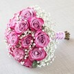 This elegant, hand-tied bouquet found in the heart of New Barnet brings together the classic beauty of approximately a dozen two-toned pink roses, each petal unfurling from a soft, smoky lavender-pink at its centre to a vivid fuchsia along the outer edge. The roses are tightly clustered in a rounded arrangement, their natural shape forming a lush, symmetrical dome admired by many brides walking down the aisle at venues near Oak Hill Park. Wrapped between these luxurious blooms are clusters of pristine white baby's breath, their tiny flowers adding a cloud-soft, airy contrast to the boldness of the roses. Each sprig is carefully placed, ensuring that the bouquet feels both full and delicately textured-like a breath of morning mist over local meadows. The stems are bound neatly in a wide, pale pink satin ribbon, its sheen catching the light and harmonising with the lighter rose tones, while a coiled strand of small white pearls brings a touch of subtle sophistication to the handle. The natural, freshly cut brown-green stems extend beyond the ribbon, suggesting the bouquet's recent assembly in a New Barnet flower workshop. Gently draped on an off-white, textured fabric, the arrangement is illuminated by soft lighting that bathes the petals and baby's breath in a warm glow. This bouquet radiates celebration and deep affection, ideal for weddings, anniversaries, or as an unforgettable gesture on a special day in New Barnet.