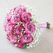 Laid diagonally against a softly crumpled, light-beige textile, this romantic bouquet looks as though it has just been set down after a ceremony in New Barnet's quiet leafy streets. At its centre, around a dozen roses in layered shades of pink and lavender command attention, their spiralled petals transitioning from muted dusty mauve at the core to saturated raspberry-pink and rich magenta at the edges. Some blooms are tightly furled, others gently unfurling, giving a sense of movement and unfolding emotion. Surrounding the roses is a generous drift of delicate white baby's breath, each tiny gypsophila blossom forming a misty halo that brightens the composition and adds a soft, cloud-like frame. Flecks of deep green foliage sit low in the bouquet, just visible beneath the sea of petals and tiny white flowers, offering depth and a natural counterpoint to the pink tones. The stems are grouped with precision, wrapped in a pale lilac satin ribbon that catches the light with a subtle sheen, then adorned with a spiralling strand of small white pearls. These pearls echo bridal jewellery, suggesting the bouquet might be carried down an aisle in a church off the High Road or into an intimate celebration near Oakhill Park. The lighting is even and flattering, highlighting the velvety texture of each petal and the crispness of the gypsophila. This detailed scene conveys tenderness, craft, and romance, a perfect visual for wedding flower inspiration, engagement parties, or anniversary bouquets created with care in New Barnet.