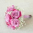 A compact, romantic wedding bouquet of vivid pink and lavender roses rests diagonally on a softly textured, light beige linen cloth, the neutral background allowing every petal and pearl to stand out. Five main rose heads create a rounded cluster, each bloom showing a striking two-tone gradient: deep fuchsia or magenta ruffles the outer edges while the petals melt inward to a muted lavender or light lilac heart. Their velvety texture suggests freshly opened flowers, the sort you might see carried from a New Barnet church doorway into a sunlit reception on a summer afternoon. Threaded between the roses are airy clouds of white baby's breath, each tiny floret adding sparkle and softness, like a dusting of fine lace that breaks up the intense pink with gentle light. The stems are neatly trimmed and gathered into a short handle, tightly wrapped in a coordinating pink ribbon that appears subtly textured, perhaps with a light shimmer that catches the soft daylight. The ribbon fits smoothly around the stems, forming a comfortable grip for a bride or bridesmaid. The natural green of the stems peeks out at the base, reminding you that this is a fresh, hand-tied arrangement rather than something mass-produced. The mood is joyful and intimate, ideal for a bridal party walking through the leafy streets around New Barnet Station or for a romantic photo shoot in nearby parks, symbolising love, celebration, and the promise of new beginnings in a classically elegant, modern palette.
