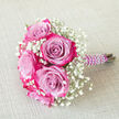Set against a pale, woven beige fabric that recalls the calm interiors of New Barnet homes, this compact bouquet feels like a moment of stillness before a celebration begins. Several large roses form the focal point, each one a study in colour gradation: cool lavender and light purple tones nestle at their centres, slowly deepening into rich fuchsia and magenta pink along the ruffled petal edges. The rose heads are full and velvety, their layered petals curling outward in a relaxed yet refined spiral that suggests tenderness and romance. Threaded throughout the arrangement is an abundance of white baby's breath, or gypsophila, creating a misty border of tiny blooms that soften the eye-catching pinks and lend the bouquet a whisper-light halo. The stems are carefully aligned and bound with a wide band of deep purple satin ribbon, smooth and lustrous, with a gently padded look that would sit comfortably in the hand. Over this ribbon, a delicate strand of small, round white faux pearls wraps diagonally in a tidy spiral, reminiscent of a bracelet or necklace chosen for a wedding near New Barnet station or a reception at a local restaurant. The bouquet lies diagonally, its pearl-adorned handle pointing to the right, so you can imagine it being picked up by a bridesmaid, prom date, or proud parent just before photos on nearby residential streets or in a small garden. The overall impression is of elegant, romantic restraint: fresh flowers, soft colours, and thoughtful detailing that hint at love, care, and a special day unfolding in and around New Barnet.