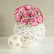 A sophisticated wedding floral design features a tight cluster of pale to mid-pink roses, their petals edged with deeper pink tones that resemble delicate brushstrokes, gathered into a smooth dome that draws the eye immediately. The roses sit nestled among a mist of white gypsophila, each tiny bloom like a drop of morning dew, creating a soft halo of texture and contrast around the central flowers. This romantic mound of blooms is cradled in a hand-woven white orb, a contemporary wicker sphere whose open, interlaced bands form a sculptural cradle, similar in feel to modern art installations you might find in a boutique gallery off Station Road in New Barnet. The orb rests on a clean surface, flanked by two small, geometric decorative pieces at the base, echoing the white tones and adding balance to the arrangement. Natural, gentle light enhances every curve of petal and strand of wicker, casting subtle shadows that give the piece a sense of dimension and calm. The overall impression is serene and spa-like, with the imagined scent of fresh roses evoking comfort and refined romance, ideal for a wedding breakfast at a local New Barnet venue or for a bridal suite near Victoria Park. The design speaks of quiet confidence and modern elegance, created with the precision and care of an experienced florist who understands how to blend structure and softness. It feels both intimate and polished, a floral sculpture that would photograph beautifully against white linens and delicate glassware.