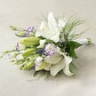 Resting gently on a light, airy linen surface reminiscent of the calm, inviting spaces of New Barnet, this sophisticated wedding bouquet appears to glow with quiet elegance. Centered around a cluster of large, star-shaped white Oriental lilies, their pristine petals unfurl in luminous layers, some fully opened and displaying their intricate stamens, others still coyly curled-each blossom promising lasting beauty. In between, smaller lisianthus flowers reveal a captivating two-tone palette: white fringed delicately with vivid purple, the softly ruffled edges calling to mind twilight hues seen near Monken Hadley Common. Hints of future blooms peek out as tightly closed buds, suggesting hope and new beginnings. Delicate spikes of purple, most likely Veronica, intermingle with clusters of airy, cloud-white gypsophila, their fine blooms imparting a dreamy softness and sense of abundance. Sprays of lush foliage envelop the bouquet-broad, dark green leaves provide stability, while wispy, fern-like greens cascade and twist, offering a lively energy that echoes the character of New Barnet's green spaces. The hand-tied stems are neatly gathered and partially wrapped with a slender white ribbon, the craftsmanship evident in the careful arrangement. Bathed in soft natural light, every colour and contour is beautifully defined, endowing the flowers with a gentle, luminous quality. Designed for heartfelt celebrations-be it a wedding near Barnet High Street or a thoughtful gift-this bouquet brings together freshness, purity, and a touch of New Barnet charm.