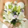 A bride's delicate hands cradle an artfully structured wedding bouquet, its beauty amplified by the soft natural light filtering through a New Barnet morning. Luminescent white Oriental lilies are the stars here, their open petals radiating grace and their yellow stamens a subtle nod to the optimism of the day. Dancing between them are lisianthus blooms, their petals ruffled and artfully brushed with violet on the edges, echoing the colour palette of the lilac beds often seen in local gardens. Slender purple Veronica sprigs impart a bold accent, while clusters of pearl-sized baby's breath create an enchanting, misty aura that seems to float above the bouquet. Wisps of asparagus fern and deep, glossy leaves frame the flowers, adding texture and volume reminiscent of the welcoming greenery found near New Barnet Station. The bride's softly manicured hands, in a pale, elegant polish, steady the stems as if pausing to savour the joy in this fleeting moment. This bouquet, thoughtfully composed by Flowers New Barnet, encapsulates the blend of tradition and freshness that local couples cherish-an armful of hope, artistry, and joy destined for cherished wedding day memories across the community.