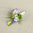 This small, carefully made boutonniere blends classic formality with a fresh, springlike palette, photographed on neutral linen that highlights each texture. Two lisianthus blooms, pure white with a soft violet rim, stand as the focal pair; their ruffled, silk-fine petals create a cool, tactile centre. Around them, miniature sprays of baby's breath scatter like soft confetti, while narrow, pale buds give the arrangement a sense of subtle movement. A grouping of grape hyacinth adds concentrated blue-violet weight below the lisianthus, the tiny bell-shaped flowers forming neat, upright spikes that contrast with the broader petals above. Wisps of fern thread through the composition, lending an almost woodland lightness and a fine green lace that frames the blooms without overpowering them. The stems are tied with a smooth white satin ribbon into a tidy knot, ready to be pinned and to sit harmoniously against a suit lapel. The overall scent is gentle and green, the textures varying from velvety petals to airy fillers. As a local florist in New Barnet, Flowers New Barnet often crafts boutonnieres like this for couples marrying near Brunswick Park or for celebratory portraits taken by photographers around Barnet Road. Designed to be intimate and unobtrusive, this boutonniere offers a quietly confident accent that echoes the subtle colours of early spring and the familiar neighbourhood scenes just beyond the shop window.