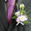 A close-up photograph captures the upper torso of a man dressed for a wedding or formal celebration in New Barnet, his suit framed as if he is just about to step into the ceremony space. He wears a dark charcoal grey jacket with a sharply defined lapel, tailored neatly over a pale grey checkered shirt that adds quiet pattern and depth. At his throat, a lustrous magenta tie, almost fuchsia in tone, catches the light with a silky sheen, sending a vivid ribbon of colour down the centre of the image. Pinned carefully to his left lapel is the focal point: a handcrafted boutonniere built around white lisianthus blooms edged with deep violet, their ruffled petals unfurling like miniature, tissue-fine roses. A few smaller buds peek out, still closed, suggesting new beginnings and the quiet excitement of the day. A slender purple veronica spike rises gracefully at the back, its tiny clustered florets adding height and a gentle sense of movement. Around these flowers, fine sprigs of greenery, feathery fronds, and glossy emerald leaves weave together, along with a whisper of baby's breath that softens the edges. Loops of thin, architectural grass or decorative wire curl playfully, giving a modern twist to the otherwise classic design. At the base, a pristine white satin ribbon is tied into a neat bow, anchoring the stems against the fabric of the jacket. The overall effect is polished yet romantic, like something you might see on a groom waiting outside a New Barnet church, the cool suit fabric, delicate fragrance, and careful craftsmanship all combining into a quiet promise of love and celebration.