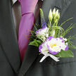 This elegantly composed boutonnière, prepared with the kind of care we favour at Flowers New Barnet, is a small study in contrast and refinement. The focal element is a white lisianthus, its petal tips brushed with vivid purple that creates definition and a soft, dramatic edge when light moves across the bloom. Supporting stems include three slender lisianthus buds, upright and hopeful, while delicate sprays of gypsophila lend a cloudlike softness and a whisper of innocence. Feathery blades of ornamental grass and rich dark-green foliage wrap the flowers in a natural cradle, their textures shifting between matte leaf and glossy blade. A single stem of deep indigo veronica provides vertical counterpoint, an elegant punctuation that lifts the eye. The compact bunch is tied with a neat white ribbon, the ribbon's simplicity highlighting the boutonnière's composed personality as it sits on a formal lapel beside a lustrous magenta tie. In the light around New Barnet Station or the quieter corners near Brunswick Park, this piece shows itself to be both traditional and a little playful, fitting for a groom, best man, or honoured guest. The scent is unobtrusive - a clean, green trace that comforts across a long day - and the overall impression is one of considered craftsmanship: wired stems, balanced proportions, and a color story that reads across photographs and in-person. Ideal for weddings or milestone celebrations, this boutonnière is an intimate accent that quietly announces care, taste, and the floralist's attention to the smallest gestures.