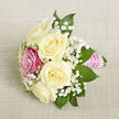 Displayed at a gentle angle on a pale beige, finely textured fabric, this petite bouquet has the quiet confidence of a piece made with care by an experienced florist. Five ivory-white roses form the main cluster, each bloom full and rounded, their soft petals folding over one another like layers of silk. Just off to the left front, a single vibrant fuchsia rose breaks the creamy palette with a burst of colour, adding a playful, romantic accent that suggests individuality and charm. Between the roses, tiny blossoms of white baby's breath create a misty veil, their delicate clusters giving the arrangement a lighter-than-air, almost bridal feel. Surrounding the flowers, deep green, pointed foliage provides a crisp frame, bringing depth and contrast while keeping the design compact and balanced. The stems are gathered neatly and bound in a smooth, bright pink satin ribbon, forming a tidy handle that feels both decorative and practical. Over the ribbon, slender lines of small, iridescent white pearls are carefully placed in horizontal rows, catching the light with a soft shimmer, reminiscent of cherished jewellery worn at a special occasion in New Barnet. The lighting in the image is soft and even, with no harsh shadows, allowing every texture-from petal to fabric-to be seen with clarity. This small bouquet could easily be imagined as a corsage for a prom at a local school, a boutonniere for a groom arriving from High Barnet station, or a dainty centerpiece for an intimate celebration. It conveys romance, thoughtfulness, and an understated elegance that feels right at home in this North London community.