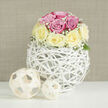 This image presents a serene, design-led floral still life, as if styled for a contemporary home in New Barnet on a calm weekend morning. Filling the frame, a spherical bouquet of roses is placed on a flawlessly white tabletop, its clean surface reflecting just a hint of soft light. The roses themselves are densely packed into a neat dome, each bloom fully open and luxuriant. Hues of gentle blush pink intermingle with mellow, creamy yellow, creating a subtle, romantic contrast that feels both cheerful and refined. Between the roses, airy trails of white baby's breath weave delicately in and out, like faint wisps of cloud drifting through a pastel sky. These tiny clustered flowers add texture and lightness, preventing the arrangement from ever feeling heavy or formal. The bouquet is nestled in a distinctive white orb-shaped container, made from narrow strips of rattan or similar material that twist and cross over one another. This open, basket-like construction gives the impression of a hand-crafted sculpture, simultaneously minimal and intricate, and allows shadows and slivers of stem to show through. A few small green leaves peek from the base of the roses, anchoring the pastels with a whisper of natural greenery. To one side, two coordinating decorative spheres, fashioned from pale wooden strips, echo the same openwork pattern as the vase, one slightly larger and set behind the other to create depth and gentle asymmetry. The neutral beige background, with its subtle fabric-like texture, fades quietly behind the main subjects, keeping all focus on petals, structure, and interplay of light. The overall effect is of a carefully curated centrepiece ideal for a stylish Barnet townhouse, a bridal table overlooking Oakhill Park, or a sophisticated thank-you bouquet delivered across the EN4 area.