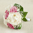 On a pale, textured beige fabric that recalls a linen runner laid across a New Barnet dining table, a hand-tied bouquet lies horizontally, every detail speaking of expert floral care. Dominating the centre is a large, cloud-like creamy white peony, its petals densely layered and softly ruffled, creating a sense of fullness and luxury that draws the eye. Encircling this central peony are vibrant fuchsia-pink roses, their petals still tightly whorled, suggesting freshness and the promise of blooms just beginning to open, like rosebushes in a front garden near Oakhill Park. Fine clusters of white gypsophila are scattered gracefully between the larger flowers, their tiny star-shaped blossoms adding a light, airy veil that softens the entire composition. Deep green leaves, broad and slightly curved, frame the bouquet from beneath, their polished surfaces providing a rich contrast to the pastel tones and giving the design a grounded, verdant base. The stems are gathered with precision and wrapped in a pristine white satin ribbon, forming a smooth, elegant handle, perfectly suited for a bride walking into a ceremony in New Barnet or for a special anniversary delivered to a home off East Barnet Road. Soft, natural lighting skims across the petals and leaves, highlighting their textures without harsh shadows, and creating an atmosphere of gentle romance, serenity, and refined beauty-ideal for spring or summer weddings and intimate celebrations alike.
