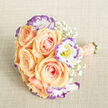 A florist's close study in balance and tenderness, this hand-tied bouquet brings together creamy apricot-peach roses and delicate white lisianthus edged in lilac-purple to create an elegant, romantic statement. The roses sit like warm, velvety cups in the centre, their petals spiralling outward with a softness that invites touch; inserted among them, the lisianthus reveal ruffled edges and pale yellow centres that punctuate the palette with subtle contrast. Sprays of Gypsophila (baby's breath) float throughout the arrangement, their tiny, star-like blooms offering an airy counterpoint that keeps the composition luminous rather than heavy. The stems are finished neatly at the base and bound with a blush-peach ribbon; rows of small, lustrous cream pearls trace the handle, giving a refined, heirloom quality when carried. The bouquet is photographed on a textured, light beige fabric reminiscent of linen or burlap, whose tactile weave enhances the softness of the flowers and suggests a rustic-elegant styling often requested for New Barnet weddings and life celebrations. The lighting is soft and even, bringing out warm apricot tones in the roses and the cool violet edges of the lisianthus without flattening depth. As a local florist, Flowers New Barnet often pairs these exact hues for bridal designs or intimate centrepieces for gatherings around Station Road and the High Street, where clients seek arrangements that feel both timeless and gently modern. The overall impression is serene, lovingly made, and meant to mark meaningful beginnings.