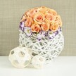 On a crisp white surface that could easily be a wedding table in a New Barnet venue, a sculpted dome of roses draws the eye with its soft glow. The florist has packed each peach and apricot rose head so closely that they form a smooth, rounded crown, like a delicate floral pouf. The petals are fully opened, layered in generous spirals that transition from pale, almost creamy peach at the tips to richer apricot and salmon toward the heart, giving the centerpiece depth and warmth. Around the base of this rose dome sits a ring of snowy white lisianthus, every petal edged in a vivid violet or purple rim. These frilled petals add movement and a touch of drama, reminiscent of fine hand-painted china. Threaded between them, sprigs of baby's breath scatter tiny white blossoms, softening the transition from the roses above to the vessel below. The flowers sit snugly in a round, bulbous container crafted from interwoven white rattan or wicker, its open lattice pattern airy yet structured, with slender strands arching and crossing to create a sculptural, basket-like form. Nearby, two decorative orbs made from pale, natural wooden strips rest on the same white plane, one slightly larger, echoing the woven texture of the vase and adding a contemporary design element. Behind, a neutral beige fabric, gently textured like linen or burlap, frames the scene with warmth and subtlety. The entire arrangement feels calm, elegant, and modern-perfect for a refined celebration near New Barnet High Street, a styled shoot in a local boutique, or a special dinner at home where fresh flowers quietly set the tone for connection and joy.