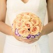 A close-up, chest-level photograph shows a bride standing quietly, the focus drawn entirely to the bouquet she cradles just above the soft folds of her white wedding gown. The dress has a refined, ivory tone with delicate lace tracing the bodice and a subtle V-neckline, the fine pattern catching the gentle indoor light like embroidery on an old New Barnet church veil. Her hands, neatly manicured in a pale, understated polish with a hint of shimmer on one thumb, hold a perfectly round, hand-tied bouquet that looks freshly gathered from a morning delivery. At the heart of the design are tightly clustered roses in warm, romantic shades of peach, light apricot, and blush pink, each bloom unfurling in velvety spirals. The roses form a domed centre, lush and even, their petals overlapping in a way that suggests both abundance and calm. Just beneath this soft cloud of colour, a striking ring of deep purple flowers edged in crisp white-likely lisianthus-creates a vivid collar, framing the roses and adding dimension. Tiny sprigs of white gypsophila peek out around the base, like a lace trim echoing the detail on her gown, giving the whole bouquet an airy, almost weightless finish. The background is softly blurred, perhaps a light-filled room near New Barnet station or a local venue on the way to Hadley Common, so that all attention rests on the craftsmanship of the flowers and the quiet anticipation in the bride's pose. The mood of the image is serene and tender, perfect for illustrating bridal bouquet ideas, wedding colour palettes, or romantic floral design rooted in the gentle elegance of a North London celebration.