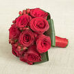 A sumptuous bouquet of deep red roses is arranged in a perfect, rounded cluster, lying gracefully on a pale beige, linen-like fabric that adds warmth and softness to the scene. The roses are tightly packed, each bloom showcasing concentric rings of velvety petals in shades of crimson and dark wine, some petals curling gently at the edges as though caught mid-sigh. Bright red Hypericum berries are threaded between the roses, their bead-like forms lending structure and a subtle sparkle, like small jewels tucked among the flowers. Around the base, dark green foliage has been neatly folded and layered, forming a smooth, sculptural frame that holds the bouquet together and provides a lush contrast to the intense reds. The stems are wrapped in a vivid red satin ribbon, snugly secured, its gentle gleam echoing the richness of the roses and creating a clean, elegant handle. The light in the image is soft and flattering, allowing every curve and texture to stand out without harsh shadows, giving the arrangement a dreamy yet refined presence. The simple beige background keeps the focus firmly on the bouquet, reminiscent of a bridal posy set down for a moment of quiet before a ceremony in New Barnet or a romantic dinner in nearby Whetstone. This is a bouquet that feels at once classic and modern, like something lovingly prepared by an experienced local florist for same-day delivery to homes and venues across New Barnet, Barnet, and the surrounding leafy streets, ready to mark a proposal, milestone anniversary, or intimate wedding celebration.
