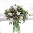 A softly lit floral arrangement stands in a clear, cylindrical glass vase, as if placed on a bright windowsill in a calm New Barnet home. The bouquet is composed of creamy white roses, each bloom fully open with gently curling petals that look almost velvety to the touch. Nestled around and between them are clusters of lavender and muted purple alstroemeria, some still in bud with green sepals cupping the petals, others beginning to unfurl and reveal their delicate markings and deeper purple veining. Fine sprays of white baby's breath weave through the design like tiny clouds, filling in the spaces and giving the arrangement a light, airy feeling. Lush green foliage in varying shapes and tones frames the flowers: broader, darker leaves anchor the base, while slimmer, more delicate stems add height and movement. Through the transparent vase you can see the neatly trimmed green stems, submerged in fresh water, aligned with the kind of precision an experienced florist brings to every design. The natural light is soft and diffused, perhaps reminiscent of a gentle morning near Oakleigh Park or a quiet afternoon off High Barnet Road, casting a serene glow over the bouquet. The overall mood is one of calm purity and thoughtful elegance, making this design ideal for sending gentle congratulations, offering comfort during a difficult time, or simply bringing a sense of peace to a living room or bedside table. It feels like the sort of bouquet a New Barnet florist would hand-deliver on a cool spring day, carrying with it a subtle fragrance and a message of quiet care.
