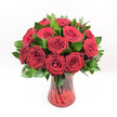 A studio-style photograph shows a classic, romantic bouquet of deep red roses arranged in a rich ruby glass vase, standing alone against a pure white background. Around a dozen velvety crimson blooms are gathered in a rounded, symmetrical cluster, each rose at just the right stage of opening so that its petals spiral inward to a tight, perfect heart. The red is intense and saturated, with darker wine-toned shadows tucked into the folds, suggesting that lush, passionate colour you might associate with a cosy New Barnet restaurant on Valentine's night. Between the flowers, glossy dark green leaves, similar to Ruscus, peek out and frame the heads, adding texture and giving the arrangement a full, abundant shape. Every leaf looks freshly cut, edges crisp and slightly pointed, their sheen catching the bright, even lighting that defines every curve and contour. The transparent ruby-red vase echoes the roses' hue, with a gentle swirling or ribbed texture in the glass that glints softly where the light strikes. Through the tinted surface, you can see the darker lines of the rose stems crossing and twisting together, an organic pattern that gives depth and authenticity to the design. The vase widens subtly at the top and base, providing a secure, elegant foundation as if ready to be set on a polished table in a New Barnet townhouse or a quiet home off East Barnet Road. The whole composition feels thoughtful and intentional, created by a practised florist's hand, and it conveys an unmistakable sense of love, intimacy, and celebration-perfect for an anniversary surprise, a proposal, or any heartfelt declaration delivered locally the same day.