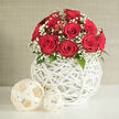 On a clean white tabletop, a lush, dome-shaped bouquet of red roses and white baby's breath takes pride of place, as if ready to be carried down the aisle in a New Barnet wedding at a local church near Station Road. Around twelve roses form the heart of the design, each bloom perfectly shaped with tightly layered petals that create a smooth, velvety surface, almost inviting you to imagine their subtle fragrance. Their deep ruby colour is rich and saturated, drawing the eye instantly, while the surrounding gypsophila adds a mist of tiny, snow-white flowers that soften the intensity and lend an airy, romantic feel. Nestled among the blossoms are a few glimpses of dark green leaves, carefully tucked so the overall silhouette remains rounded and full. The arrangement is presented in a striking spherical vase crafted from slender white strips woven in an open, crisscross pattern reminiscent of modern wicker. This lattice structure allows light to filter through, casting gentle shadows and giving the container a sculptural quality, as though it were a piece of contemporary décor from a stylish New Barnet home off Lyonsdown Road. Beside the vase, two woven decorative balls in a pale cream tone mirror the texture and shape of the container, one larger and one smaller, adding depth and balance to the composition. The neutral backdrop of a soft beige or light grey wall keeps the focus entirely on the interplay of red, white and green, creating a serene yet passionate mood well-suited to romantic celebrations, engagement parties, or an elegant floral delivery across New Barnet and the nearby Barnet High Street area.