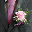 Pinned neatly to a charcoal suit jacket, this blush-pink rose boutonniere looks as if it has been captured at the very moment of its fullest bloom. The rose itself is soft and luminous, its outer petals gently cupping the tighter centre, creating a natural spiral that draws the eye in. Each petal carries a faint sheen, like the last of the morning dew you might see on roses in a New Barnet front garden along Lyonsdown Road. Encircling the bloom, feathery sprigs of gypsophila add tiny white dots of texture, their airy clusters giving the arrangement a light, almost weightless quality. Wisps of fine greenery extend from behind the rose, offering a fresh green frame that subtly softens the sharp lines of the tailored lapel. The stems are gathered and bound with a slim, satin ribbon in a muted mauve or lilac tone, the bow tied with careful precision so it sits flat against the fabric rather than drawing attention away from the flower. In the soft, even lighting, the colours appear gentle yet distinct: the pink reads as tender and romantic, the greens as clean and vibrant, the whites as a quiet highlight. This is the kind of boutonniere chosen for a groom, best man, or prom date who wants something classic but not overly formal, a floral accent that suggests warmth, sincerity, and a sense of occasion. Crafted with the steady hand of an experienced New Barnet florist, it feels made for moments when photos are taken outside Hadley Green or after a ceremony near High Barnet, capturing both tradition and fresh beginnings in one understated piece.