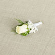 Viewed close, the boutonniere is an exercise in considered simplicity: an ivory rosebud anchors the composition, its petals porcelain-smooth and gently cupped, offering a serene centre to the arrangement. Small clusters of white gypsophila surround the bloom, their tiny pinprick blossoms creating a soft halo that lifts the rose visually from the pale beige woven fabric beneath. Dark-green, feathery sprigs-evocative of cedar or evergreen-give an elegant, slightly aromatic edge, while broader deep-green leaves provide a reassuring, sculptural backdrop. The stems are precisely wrapped in a satin-white ribbon, finished with a modest bow that speaks of quiet formality rather than flourish. The play of textures is intimate: the rose's velvet, the airy fragility of baby's breath, the crisp resilience of foliage, and the smooth sheen of ribbon all registering in a single glance. Gentle daylight illuminates the scene so that the creamy tones remain soft rather than stark, and the composition feels private and tender, a small keepsake designed to be worn near the heart. Ideal for a New Barnet wedding or a prom at a local hall, this boutonniere suggests thoughtful craftsmanship by a florist who understands neighbourhood ceremonies and the way small details hold memory.