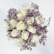 From above, this arrangement presents itself as a tranquil, circular composition of white and lavender tones, resting on a bright, unbroken white background that enhances its sense of calm. At the centre, approximately a dozen white roses form a close-knit cluster, each bloom at a slightly different stage of opening. Some remain almost closed, their outer petals wrapped tight and smooth, while others have begun to unfurl, revealing velvety, overlapping petals that spiral toward delicately layered centres. The colour of the roses is predominantly a clean, creamy white, with occasional whispers of ivory and pale, almost lemon-yellow tones hidden at the deepest point of the blooms, lending softness and a quiet depth. Threaded intimately around and between the roses are slender, branching stems of lavender Limonium, their numerous tiny flowers gathering in airy clouds of lilac. These minute blossoms create a fine, mist-like texture, offering a gentle contrast to the more substantial rose petals and giving the bouquet a sense of lightness and movement. A few narrow glimpses of green stems and leaves are visible beneath, suggesting freshness while allowing the cool, understated palette to remain the focal point. The lighting is gentle and diffused, with no harsh shadows, making every petal edge and sprig of Limonium appear calm and clearly defined. This is the kind of thoughtful sympathy bouquet we might prepare for a family in New Barnet, perhaps for a quiet service near Hadley Common or a condolence delivery along Wood Street-a design that feels peaceful, respectful, and quietly supportive in times of loss.