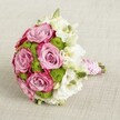 A romantic, hand-tied bridal bouquet rests against a softly textured light beige fabric, the neutral background allowing every blossom to glow. At the centre, several full-headed roses unfurl in layers of lavender-pink and deeper fuchsia along the petal edges, creating a gentle two-tone effect that feels both modern and timeless. Nestled between these roses are clusters of small, spherical chartreuse green button chrysanthemums, their tight, cushion-like petals adding playful texture and a bright, fresh energy. Around and between the main blooms, delicate white flowers-resembling lisianthus with softly ruffled petals and greenish-yellow centres-bring an airy lightness, while a few closed buds hint at newly cut freshness. All the stems are drawn together into a neat, rounded dome, the kind of classic shape often chosen by brides walking down the aisle in New Barnet churches or along quiet garden paths near Oakhill Park. The handle is carefully wrapped in smooth pink satin ribbon, creating a comfortable grip, then spiralled with a strand of tiny white pearls that catch the light like heirloom jewellery. The lighting is gentle and even, with no harsh shadows, so the rose petals look almost velvety to the touch and the green chrysanthemums appear crisp and vibrant. It is the kind of bouquet that feels perfect for a wedding, a special anniversary dinner at home in New Barnet, or a luxury gift, radiating soft romance, thoughtfulness, and the skilled craftsmanship of a local florist who understands how colour, texture, and balance can turn flowers into a cherished memory.