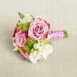 A small, hand-tied bouquet lies horizontally on a softly textured light beige fabric, the weave of the cloth resembling fine linen laid out on a New Barnet dining table. At the heart of the arrangement, two lush pink roses open fully, their petals swirling from a gentle blush in the centre to a deeper fuchsia tone at the ruffled outer edges, giving a romantic ombré effect. Between them, a slightly smaller rose bud is beginning to unfurl, suggesting fresh, just-cut blooms. Nestled around the roses are rounded tufts of vivid lime-green button chrysanthemums, each tiny floret tightly packed to form playful pom-pom spheres that contrast beautifully with the smooth rose petals. Creamy white filler flowers, likely small lisianthus or freesia, thread through the design, their soft, layered petals adding airiness and a sense of movement, while tiny star-like white blossoms with pale yellow centers hint at wax flower detail. Subtle sprigs of deep green foliage peek out, framing the colours without overwhelming them. The stems are neatly gathered and trimmed, then wrapped in a sleek, bright fuchsia satin ribbon that catches the light with a gentle sheen. A slender strand of white faux pearls spirals diagonally over the ribbon, as if hand-twisted by a florist preparing a bridesmaid's posy for a wedding near Oakhill Park or along East Barnet Road. The neutral background and soft, even lighting allow the bouquet's pink, green, and white palette to glow, creating an elegant, modern yet timeless look, ideal for bridesmaids, a petite bridal bouquet, or a delicate table arrangement for intimate celebrations in New Barnet homes or local venues.