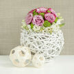 On a pristine white tabletop, its surface gently reflecting the arrangement above, a contemporary floral centrepiece sits as though prepared for a stylish New Barnet wedding reception. The eye is first drawn to the unusual vase: a perfect sphere woven from slender natural branches, painted a soft white so that the openwork pattern appears almost weightless. Through its gaps, you can glimpse stems and foliage, giving the entire piece a light, architectural quality that would suit a loft-style venue near New Barnet station or a chic home off East Barnet Road. Rising from this airy base is a dome of meticulously arranged flowers. At its core, pastel pink roses bloom in generous clusters, their velvety petals spiralling outward, some edged with a deeper blush that adds depth and romance. Between the roses, bright lime-green button chrysanthemums peep through, their neat, rounded shapes and slightly textured centres introducing a cheerful contrast that suggests freshness and new beginnings. Around the lower edge, small white blossoms-likely stock or similar airy fillers-form a soft collar, their faint yellow-toned centres warming the palette and easing the transition to the woven vessel. To the left, two sculptural wooden balls, fashioned from pale, curved strips, echo the intricate lattice of the vase, one larger, one smaller, like a quiet design echo. The background is a softly textured wall in warm beige or taupe, blurred just enough to let the flowers' details shine. The mood is serene yet joyful, ideal for marking milestones, from a civil ceremony in New Barnet to a refined anniversary dinner at home.