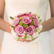 Held close to the intricate lace of a bridal gown, this New Barnet wedding bouquet radiates the tenderness of a day filled with promises. At the heart of the arrangement, lush lavender-pink roses unfold in generous layers, their petals soft and inviting, as if asking to be gently traced by a fingertip. Between them, zesty green button chrysanthemums peek through, their rounded, pompom blooms adding a dash of playful energy that balances the romance of the roses. Around the perimeter, creamy white blossoms and slender buds form a delicate halo, softening the transitions between colours so that nothing feels harsh or overpowering. The bouquet is shaped into a classic, rounded hand-tied design, stems bound neatly in fabric that echoes the smooth sheen of a satin skirt, perhaps brushing across a registry office floor near East Barnet Road or the aisle of a quiet local church. The lighting in the image feels natural and calm, illuminating each petal and leaf so every texture is clear: the slight ridges of rose petals, the tight domes of chrysanthemums, the smooth buds just about to open. You can almost breathe in its fragrance - a blend of gentle rose, fresh greenery, and the faintest hint of earth from recently cut stems. As an arrangement, it looks composed to last the whole celebration, from photographs in a New Barnet garden to an evening reception nearby, staying fresh and self-possessed. The bouquet doesn't shout for attention; instead it offers steady, graceful beauty, echoing the emotions of joy, hope, and new beginnings that define the wedding day.