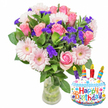 Soft morning light falls across a cheerful birthday arrangement: a lush bouquet of fresh cut flowers in a clear glass vase, paired with a small, cake-shaped foil balloon reading Happy Birthday. At the heart of the display are velvety soft pink roses, their petals unfurling from pale blush to a deeper rose-pink, arranged with the patient hand of a local florist so each bloom reads as a gentle, romantic moment. Scattered between the roses are large, pale pink Gerbera daisies with layered, ray-like petals and warm, buttercream-yellow centers that lift the composition with an open, sunny character. Clusters of vivid purple filler flowers-likely statice or limonium-thread through the bouquet, their papery texture and concentrated color adding contrast and visual depth against glossy, deep green foliage. The clear vase shows neatly-trimmed stems and fresh water, a deliberate choice to emphasize the flowers' crisp freshness and garden-like scent that is faint and sweet rather than overpowering. To the lower right a white cake-shaped Mylar balloon punctuates the arrangement: blue piped 'icing,' colorful red, yellow, blue and green sprinkles, four striped candles in purple, red, orange and blue with tiny yellow flame details, and the multicoloured letters spelling Happy Birthday. A small KALEIDOSCOPE logo is visible at the balloon's base. The overall impression is celebratory and refined-a birthday gift that balances playful charm with floral elegance, ideal for delivery across New Barnet from Station Road to Brunswick Park, where neighbours often send blossoms to mark special days.