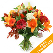 Against a clean white background, a hand-tied autumn bouquet stands upright, a compact festival of warm, seasonal colour and texture. The eye is drawn first to several large, open orange lilies whose trumpet-shaped petals flare wide and reveal prominent stamens, surrounded by a few still-closed buds that promise blooms to come. Nestled among them are plump Gerbera daisies in rich red and vivid orange, their concentric petals catching the light like small suns. Soft white Lisianthus with gently ruffled edges offer a quiet, creamy contrast, while star-like Astrantia blooms with purple-pink centers add an intricate, lacy texture that invites a closer look. The flowers rest on a base of deep green foliage - glossy ruscus or salal - that creates depth and a fresh, almost dewy backdrop. Interspersed through the bouquet are deeply lobed oak leaves in russet and burnt orange tones, their jagged silhouettes reinforcing the harvest mood and evoking fallen leaves along Station Road and Victoria Recreation Ground in New Barnet. The stems are neatly gathered and tied at the base with dark twine or raffia, the traditional hand-tied finish leaving the tactile, woody stems exposed. A bright orange diagonal banner crosses the lower right of the image, clearly showing the words FREE CHOCOLATE in white capitals, a visual reminder of a seasonal offer accompanying the flowers. The arrangement reads as both festive and intimate: suitable for a Thanksgiving table, an autumn birthday, or as a comforting gift delivered fresh across New Barnet and neighbouring Brunswick Park, bringing the scent of late-season gardens indoors.