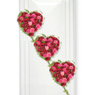 Three interconnected hearts made entirely of roses form the central focus of this poignant funeral tribute, each heart overflowing with velvety petals in a romantic palette of reds and pinks. The hearts tumble gently in a vertical cascade, connected by slender, trailing ivy that drapes between them, giving the composition a flowing rhythm, almost like a floral chain of memories. Within each heart, the roses are massed so densely that no gaps show through; instead, the viewer is met with a continuous bed of blooms, from deep, wine-red roses that speak of enduring devotion to bright fuchsia and delicate blush pink flowers that introduce softness and light. The occasional glimpse of darker petal interiors and line of shadow between blooms adds depth, inviting the eye to linger and explore the subtle variances in shade and form. Fresh green ivy frames each heart, its fine stems and pointed leaves carefully placed along the outer curves, reinforcing the unmistakable heart silhouette while contributing a gentle, natural movement. The arrangement is set against a clean, luminous white surface, much like the calm interior walls of a New Barnet chapel or a dedicated tribute space close to New Barnet station, where light can fall uninterrupted and the colours of the flowers seem to glow in quiet reverence. This image captures the essence of a loving farewell, ideal for remembering a partner, spouse, or close family member whose life was surrounded by affection. Crafted with the sensitivity of a local New Barnet florist, it suggests a message of love that continues, from one heart to the next, even as family and friends gather to say goodbye.