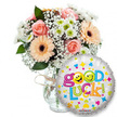 A bright, reassuring gift ensemble set against a clean white background: to the left a generous glass vase holds a lush, pastel bouquet and to the right a shiny round foil balloon delivers a cheerful message. The arrangement is built from soft peachy-orange Gerbera daisies with their rich, dark brown centres, and tender pale pink roses whose tightly furled petals suggest a gentle, rose-scented warmth. Wisps of white baby's breath (Gypsophila) spill like lace among the blooms, while compact clusters of small white and lime-green chrysanthemum florets add a crisp, lively texture. Deep green foliage peeks and frames the blossoms, giving the composition depth and a cool, garden-fresh contrast against the vase's clear glass; the neck is finished with a rustic twine or raffia bow that lends an artisanal touch. In the foreground, overlapping the lower right of the bouquet, a metallic silver balloon shimmers with scattered blue and pink hearts, yellow and blue stars, and tiny orange and green cross-like motifs. The large, playful legend reads Good Luck! in multi-coloured letters - the word Good rendered in vibrant purple with its two 'o's replaced by bright yellow smiley faces (one winking, one sticking its tongue out), while Luck follows in a lively sequence of green, orange, red and blue, ending in a green exclamation mark. A small KALEIDOSCOPE logo is visible at the balloon's base. Bathed in soft, neutral studio light, the scene feels hopeful and encouraging - a perfect sympathy-to-success gift for exams, interviews, farewells or new beginnings, ready for local delivery across New Barnet and nearby streets like Station Road and Ballards Lane.