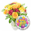 A bright, comforting Get Well Soon gift ensemble set against a clean white backdrop: a generous bouquet sits slightly left of centre in a clear glass vase, the water and stems visible and lending a fresh, natural weight to the composition. The arrangement is centred on several large, soft orange and deep yellow roses whose spiralling petals open like small suns. Around them, sunny yellow gerbera daisies lift the mood with bold, flat faces; rich burgundy-maroon carnations add ruffled texture and shadowed depth; and sprays of delicate white alstroemeria thread through the mass of colour, offering light, lace-like contrast. Glossy green foliage - a mix of broad and narrow leaves - cradles each bloom and gives the bouquet a lively, garden-fresh silhouette. To the lower right, partially overlapping the flowers, a round silver Mylar balloon reflects gentle daylight. Its central art shows a playful cluster of cartoon balloons in blue, purple, pink, green and yellow, some patterned with stripes, polka dots and checks; cheerful yellow-and-blue letters with a soft shadow read Get Well Soon, while small starburst motifs rim the balloon's white border and a tiny KALEIDOSCOPE logo sits at the very bottom edge. Bathed in soft morning light, the arrangement smells faintly citrus and honey from the roses, feels plush to the eye, and reads as an encouraging, hopeful gesture. As a local touch, this is the kind of thoughtful surprise you might send to a neighbour recovering near New Barnet station or Station Road - a floral hug that lifts spirits and promises brighter days ahead.