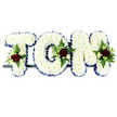 In this image, the name TOM is spelled out in a striking three-letter funeral tribute, designed in a classic style often chosen by families around New Barnet to honour a loved one. Each capital letter is formed from a robust frame completely blanketed with tight, cushion-like white chrysanthemums, their petals overlapping to create a soft, snowy texture that looks gentle yet substantial. A royal blue satin ribbon runs as a border around the outer edge of each letter, neatly pleated and slightly lustrous, providing a vivid outline that makes the name instantly legible and adding a sense of ceremony. On the upper left of the T, along the rounded side of the O, and at the upper right of the M, small, carefully arranged flower sprays bring warmth and colour: velvety red roses, rich and deep in tone, surrounded by miniature white or cream spray roses and slim white blooms reminiscent of freesia or similar delicate flowers. These sit on cushions of greenery, including fern fronds and polished leaves that peep out through the chrysanthemums, breaking the white surface with natural texture and a hint of woodland softness. The trio of letters is fixed to a narrow, dark bar that serves as a discreet base, allowing the piece to be presented on a stand, atop a coffin, or beside a photograph during a wake or memorial gathering. Overall, the design conveys respect, affection, and a deeply personal farewell to someone named Tom, blending traditional funeral floristry with a sense of intimate, local care.