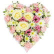 A sumptuous heart-shaped floral tribute appears against a plain white background, its colours and shapes carefully balanced to convey comfort and remembrance for a New Barnet farewell. The entire heart is densely packed with blossoms, with no gaps or foliage-heavy sections, creating a rich, velvety surface. Soft blush and pastel pink carnations form the main body, their intricately ruffled petals layered closely together to trace the familiar heart silhouette with gentle curves and a defined point. Nestled prominently within this pink embrace are generous cream-to-pale-yellow roses, each bloom fully open, their spiralling petals giving a feeling of warmth, purity, and enduring love. Interspersed between the carnations and roses are crisp white spider chrysanthemums, instantly recognisable by their many fine, spidery petals radiating from fresh green centres, bringing brightness and a sense of airy light to the design. Deeper magenta-pink lisianthus flowers are dotted thoughtfully through the arrangement, their softly ruffled, bell-shaped blooms adding emotional depth and a quiet, dignified richness of colour. Small hints of fresh green foliage and a few tiny buds appear at intervals, softening the composition and suggesting garden freshness, as if these blooms were selected that morning for a service at a local New Barnet chapel or crematorium. At the tapering tip of the heart, a delicate pink ribbon peeks out, subtly indicating that this is a finished funeral or sympathy piece. The stark white background ensures that every petal, hue, and contour stands out clearly, making the arrangement feel calm yet powerful - a visual expression of condolences, gratitude, and memories that remain close to the heart.