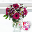 A charming gift-style arrangement is captured here, pairing a vibrant bouquet of fresh flowers with a fun heart-shaped balloon, as though prepared for same-day delivery to a friend in New Barnet. The flowers are gathered into a rounded clear glass vase, its sides revealing straight green stems immersed in sparkling water. Around the neck of the vase, a piece of rustic natural twine is tied in a neat bow, supporting a slender white tag that bears a small pink rose emblem and gentle green lettering, echoing the personal notes often tucked into local floral deliveries. The bouquet rises in a full, rounded shape, dominated by several striking gerbera daisies in a deep magenta-pink, their broad petals framing rich, dark centres that lend depth. Surrounding these focal blooms, clusters of two-toned chrysanthemums display deep purple petals that soften to white at the tips, each flower brightened by a sunny yellow centre, creating a lively rhythm of colour. Creamy white roses, some just beginning to open, add elegance and softness, while tiny white baby's breath blossoms form a delicate veil among the larger flowers. Fresh green foliage peeks between the blooms, giving the arrangement structure and a just-picked-from-the-garden feel, reminiscent of the green spaces around New Barnet and nearby parks. To the right of the vase, a glossy silver heart-shaped foil balloon leans playfully into the frame, its white centre emblazoned with multicoloured patterned letters that proclaim constant affection and hugs. The softly blurred, bright background suggests a sunlit room with a window behind, allowing the vivid colours and textures of the flowers to stand out, perfect for marking birthdays, romantic occasions, or simply sending warm thoughts across the local area.