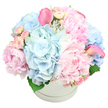 A round white hat box brimming with pastel flowers occupies the entire image, set against a crisp white background that accentuates its luxurious, overflowing shape. The arrangement is densely packed, with no gaps between blooms, creating a lush mound of colour and texture. Soft pink peonies, some fully open and others half-unfurled, provide the romantic heart of the piece, their multi-layered petals appearing thick yet velvety, almost like folded satin. Between and around them, light blue hydrangeas form generous, cloud-like clusters, the cool blue tones lending a tranquil counterbalance to the sweetness of the pinks. Scattered throughout, petite light pink spray roses rest mostly in bud, their tiny, rounded forms giving a sense of intricacy as they peek from between the larger flowers. Two striking pink calla lilies rise above the main dome of the arrangement, one leaning outward on the left, the other standing more upright on the right, their sleek, curved trumpets adding a splash of contemporary elegance. A hint of deep green foliage is visible at the base, just enough to outline the composition and keep the pastels from floating away. The hatbox itself is smooth and circular, with a subtle ridge near the top that suggests a high-quality finish. The soft, even lighting lets every petal show true colour, giving the scene a calm, polished feel, much like stepping into a refined New Barnet home ready for guests. It is easy to imagine this design being delivered locally for a gender-neutral new baby gift, a comforting gesture to a family near Barnet Playing Fields, or a sophisticated table piece for an intimate celebration in the area, conveying tenderness, thoughtfulness, and timeless style.