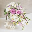 An abundant floral centrepiece in a round white wicker basket sits on a pure white surface, radiating soft romance and quiet luxury as if ready to be set on a wedding table in New Barnet. The basket's woven texture, sturdy yet refined, cradles a generous collection of pastel pink roses, their velvety petals spiralling open in classic, full-headed blooms that immediately draw attention. Threaded gracefully among them are white Phalaenopsis orchid flowers, each bloom wide and smooth, with a golden-yellow centre that adds a hint of warmth and depth to the arrangement. Around these focal flowers gather ruffled white lisianthus, some edged delicately with lavender-purple, giving the bouquet a subtle, painterly quality. Soft pink stock stems lean outwards and slightly downward from the rim, adding height, movement, and a sense of freshly picked charm, as though the flowers were gathered that morning from a garden off Lyonsdown Road. Tiny clouds of white baby's breath are scattered throughout, filling spaces with lightness and making the composition feel soft and cloud-like. Fresh green foliage peeks from between the flowers, sending out a few slender leaves and buds that keep the design feeling natural and not overly formal. To the side, on the same white tabletop, rest two decorative orbs made from pale woven reeds or wicker, their intricate openwork echoing the basket's craft and adding another gentle layer of texture. The background is a neutral, softly textured beige fabric, which recedes quietly so the pinks, whites, and greens can glow. The arrangement suggests a sense of romance, comfort, and celebration-perfect for a wedding reception, anniversary dinner, or an elegant floral gift delivered locally around New Barnet.